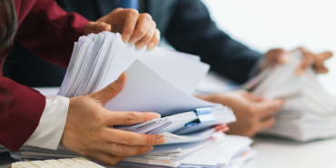 Two businesswomen are working together to search through important documents from a pile of documents on a table to verify the authenticity of the documents that were previously signed in a contract.