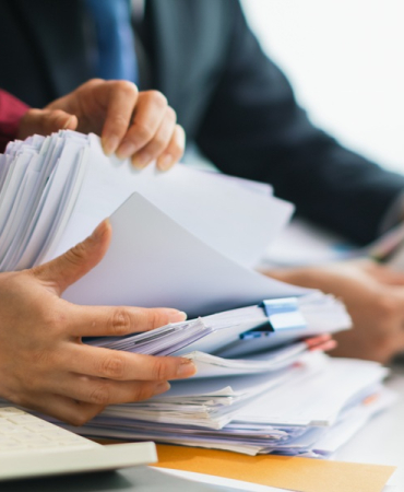 Two businesswomen are working together to search through important documents from a pile of documents on a table to verify the authenticity of the documents that were previously signed in a contract.