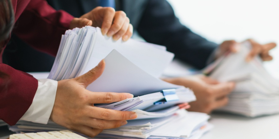 Two businesswomen are working together to search through important documents from a pile of documents on a table to verify the authenticity of the documents that were previously signed in a contract.